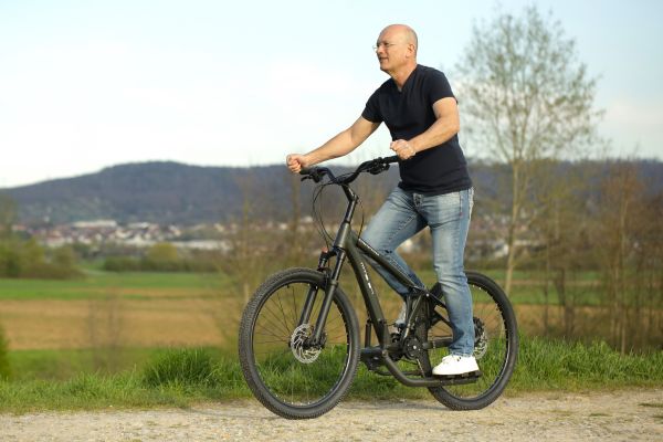 Mann steht auf einem Fahrrad ohne Sattel auf einem Schotterweg in hügeliger Landschaft.